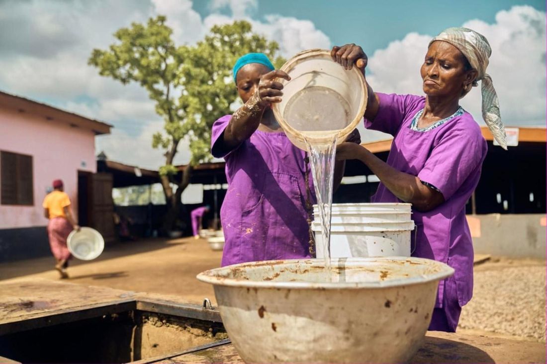Two women pour clean water from a basin into a bucket in a sunny village courtyard